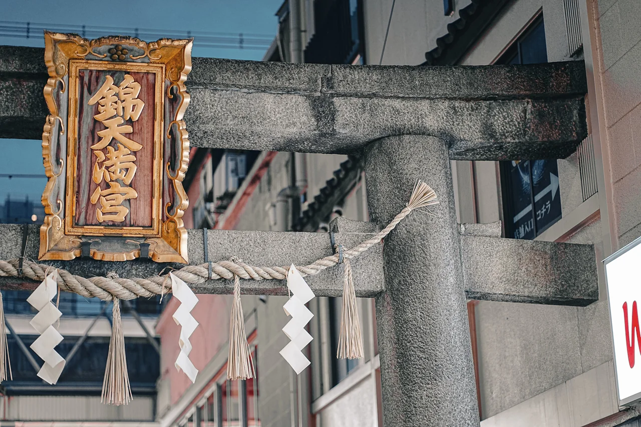 京都錦天滿宮石造鳥居上方被現代建築物穿過,黃金牌匾與白紙符掛於繩上